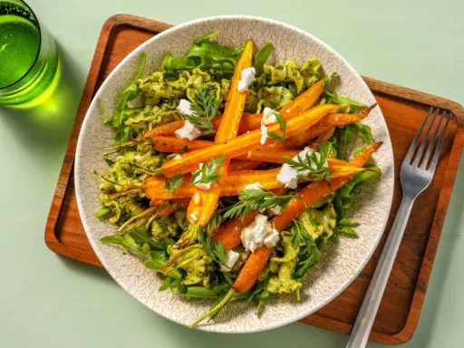 Un plat de pâtes vertes garni de carottes rôties et de fromage frais, avec des fanes de carottes pour la décoration.