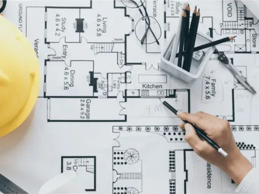 Un arquitecto o proyectista de una obra revisa planos detallados de una casa, con casco amarillo, lápices y calculadora.