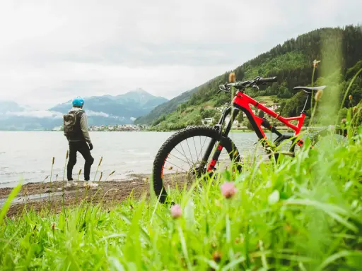 En person med hjälm och ryggsäck står vid en sjö med en mountainbike framför sig. Berg och en stad syns i bakgrunden. Perfekt för att cykla MTB.