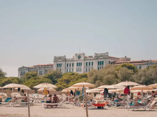 Entspannte Menschen liegen unter Sonnenschirmen am Lido di Venezia Beach. Im Hintergrund thront ein imposantes Gebäude vor üppigem Grün.