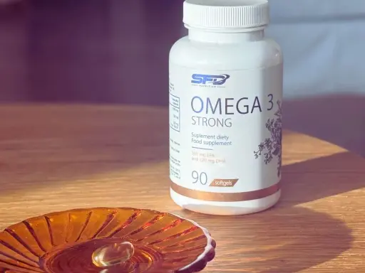 Biała butelka suplementu diety Omega 3 Strong. Na małej, bursztynowej podstawce leży jedna kapsułka. Kwasy omega 3 na co? Na zdrowie!