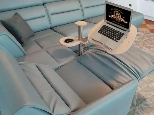 Comodo tavolino porta pc in legno e metallo, agganciato a un divano in pelle azzurra, con laptop aperto e mouse.