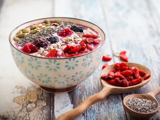 Miseczka smoothie z owocami, nasionami chia, pestkami dyni i słonecznika. Obok łyżka z jagodami goji. Nasiona chia jak jeść? Dodaj do smoothie!