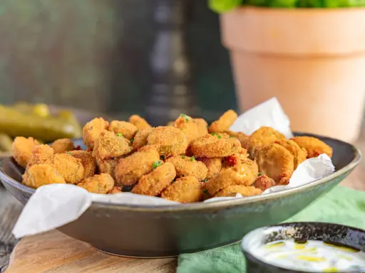 Knusprig frittierte Gurkenscheiben, **fried pickles**, in einer Schale, daneben ein Dip und ganze Gewürzgurken.