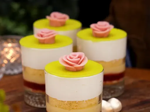Fyra små prinsesstårta i glas, dekorerade med rosa marsipanrosor och gul marsipanlock. Vackert uppdukat med ljus i bakgrunden.