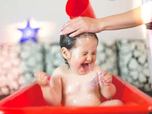 Ein Baby lacht und schreit vor Freude, während seine Haare gewaschen werden. Wasser spritzt, und es hält ein lila Band.