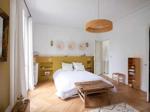 Une chambre lumineuse avec une tête de lit peinture jaune moutarde, des décorations murales en paille et un lit blanc douillet.