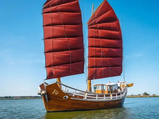 Ein hölzernes Dschunke Schiff mit roten Segeln gleitet auf ruhigem Wasser unter blauem Himmel.
