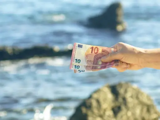 Dłoń trzyma banknoty euro na tle morza. W Czarnogórze obowiązuje euro, więc te pieniądze przydadzą się na wakacjach.