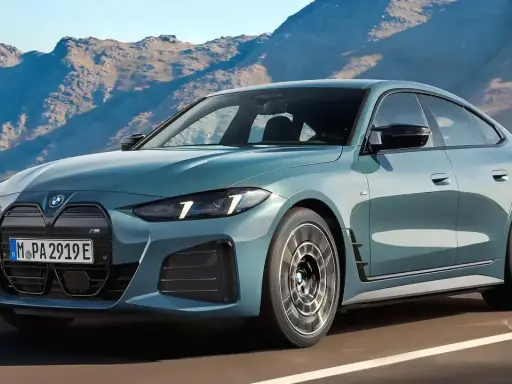 Der neue BMW i4 Facelift in einem eleganten Blau fährt auf einer Küstenstraße vor einer Bergkulisse.