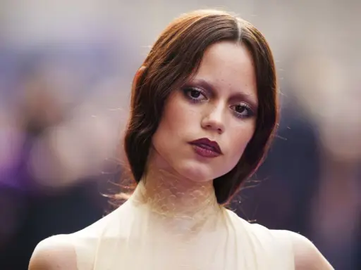 A young woman with auburn hair and a dramatic, dark lipstick looks directly at the camera. Her eye makeup is smudged, giving her a tired girl makeup look, as if she's been out all night.