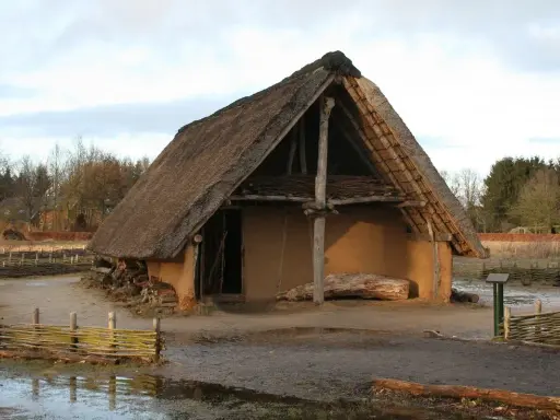 Ein rustikales Bauernhaus aus dem Mittelalter mit Strohdach und Lehmwänden, umgeben von Holzzäunen und Bäumen.