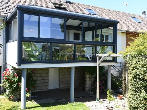 Moderne veranda adossée au mur, avec une chaise verte visible à travers les grandes baies vitrées. Le soleil brille sur le jardin verdoyant.