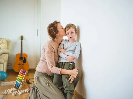 Madre besa a su niño independiente, apoyado en la pared. Juguetes y guitarra en el fondo.