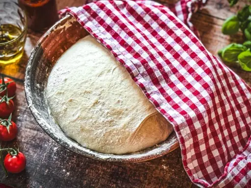 Frischer Pizzateig ruht in einer Schüssel, bedeckt mit einem rot-weiß karierten Tuch. Die Frage: wie viel Hefe in Pizzateig?
