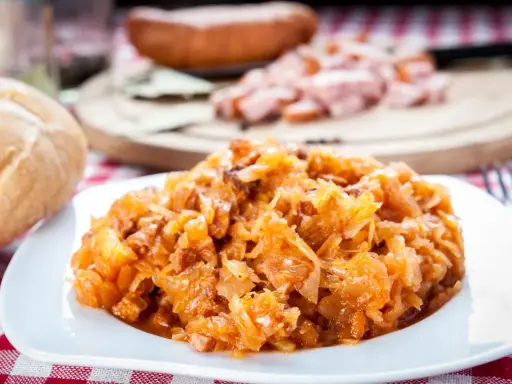 Pyszny bigos Ani, gotowany z pasją, na talerzu. Obok bułka i kawałki kiełbasy.