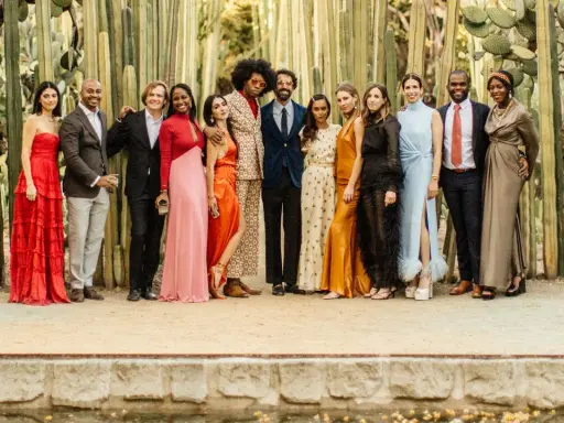 A diverse group of people pose in front of tall cacti, showcasing a vibrant summer color palette with dresses in red, pink, orange, and gold, alongside suits in blues and browns.