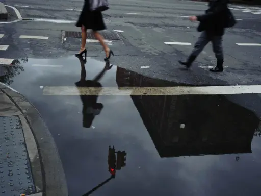 Jay Maisel photography captures a rainy street scene with blurred figures crossing, their reflections distorted in puddles, mirroring the city's mood.