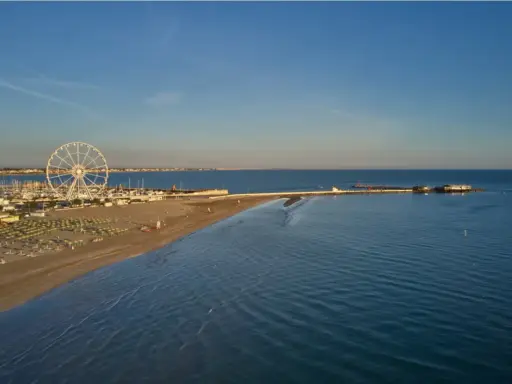Vista aerea di una spiaggia di Rimini con ruota panoramica, porto turistico e mare calmo sotto un cielo azzurro.