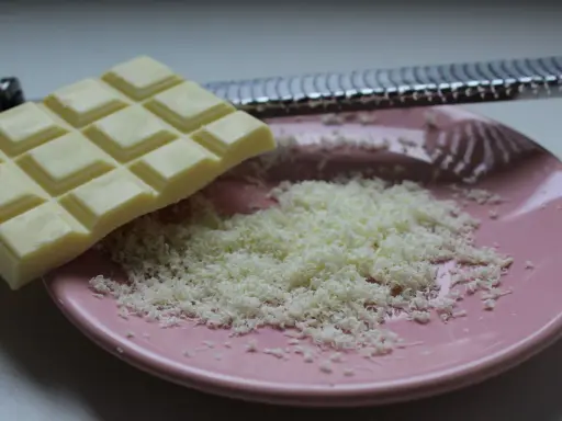 Une râpe à fromage argentée est posée sur une assiette rose, recouverte de copeaux de chocolat blanc fraîchement râpé.