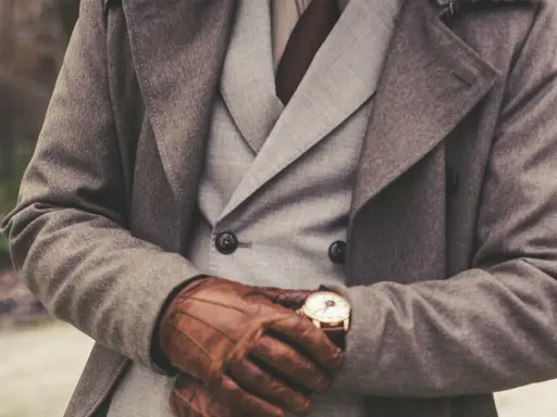 Elegancki mężczyzna w płaszczu, kamizelce i skórzanych rękawiczkach, sprawdzający zegarek. To uosobienie stylu, prawdziwy dandys kto to.