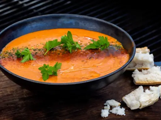 En skål med krämig, orange soppa, troligen en smakrik mojo rojo, garnerad med persilja och serverad med brödbitar.