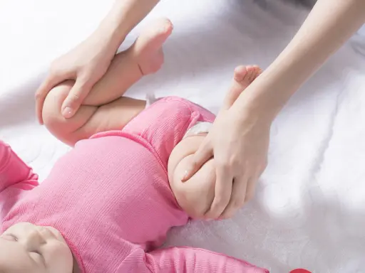 Delikatne ruchy dłoni pomagają w diagnostyce dysplazji stawu biodrowego u niemowlaka w różowym body.