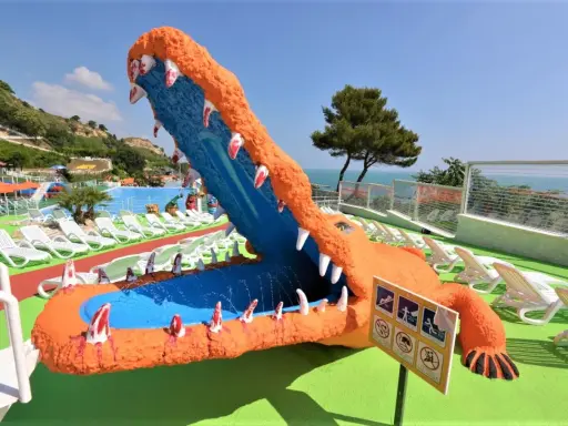 Un gigantesco coccodrillo arancione con la bocca spalancata, un divertente scivolo acquatico, si affaccia su una piscina e lettini bianchi, con una rigogliosa riva verde fermo sullo sfondo.