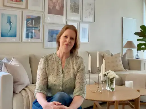 En kvinna sitter i en ljus soffa omgiven av olika inredningsstilar. Tavlor med havstema och en stor grön växt bidrar till den lugna atmosfären.