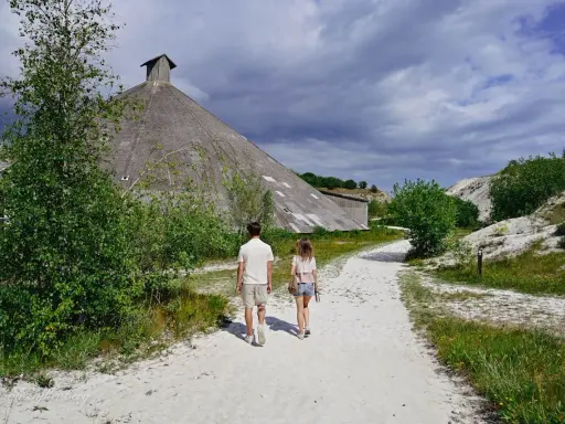 Två personer går på en grusväg mot en stor, konformad byggnad. Omgivningen påminner om vita klippor i Danmark, med grönska och en dramatisk himmel.