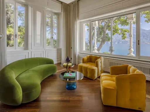 Salotto con divano verde curvo e poltrone gialle, tavolini di design e vista lago. Un'elegante casa Art Déco.