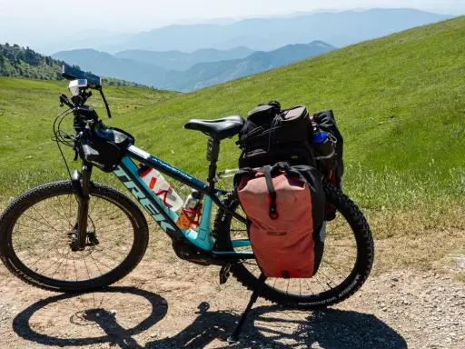 Bereit für e bike touren: Ein Trek E-Bike mit Gepäcktaschen steht auf einem Schotterweg, umgeben von grünen Hügeln und Bergketten.