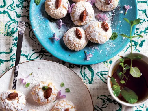 Biscotti al cocco spolverati di zucchero a velo, guarniti con mandorle e fiori viola, accanto a una tazza di tè alla menta.