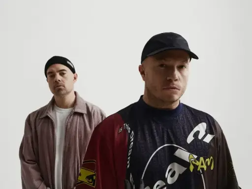 Dwóch mężczyzn, jeden w czapce beanie, drugi w czapce z daszkiem. Na koszulce jednego z nich widoczny jest **guzior fala tekst**.