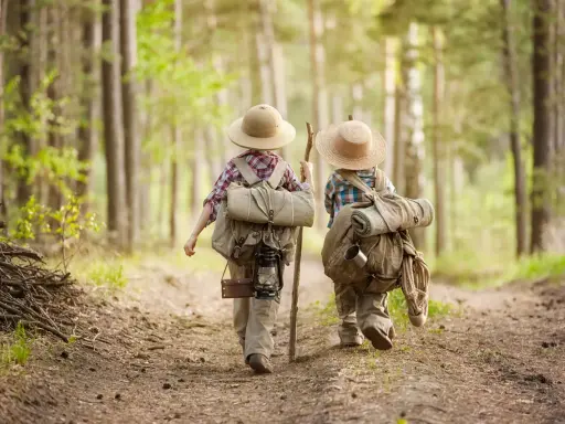 Dwoje dzieci w kapeluszach i z plecakami wyrusza na leśną ścieżkę. To przygoda i nauka survivalu dla dzieci.