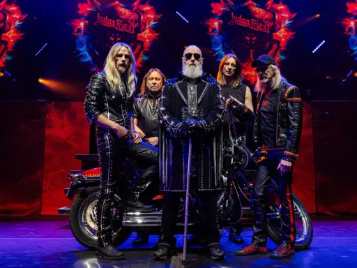 Medlemmar av Judas Priest poserar på scenen med en motorcykel. Bandets logotyp syns i bakgrunden.