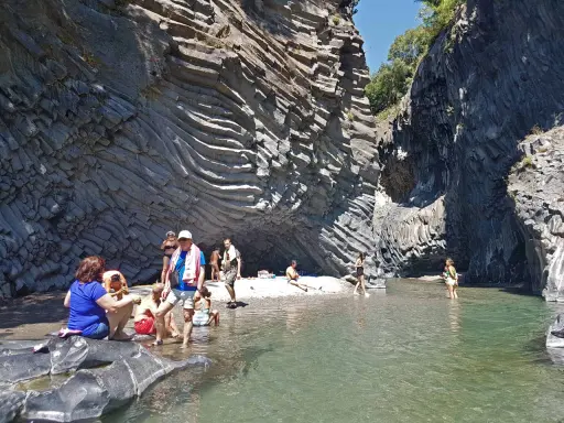 Persone si rilassano e si rinfrescano nelle acque cristalline delle cascate dell'Alcantara, circondate da imponenti pareti rocciose basaltiche.