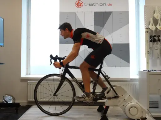 Mann auf einem Rennrad-Trainer, der sein Lungenvolumen trainiert. Hinter ihm eine Wand mit Triathlon-Logo und Messmarkierungen.