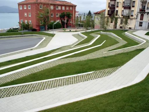 Diseño moderno con césped y caminos de hormigón que forman patrones ondulados. El suelo drenante permite el paso del agua, integrándose con el entorno urbano y el mar al fondo.