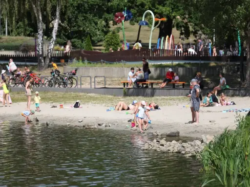 Rodzina odpoczywa nad wodą na piaszczystej plaży Jeziora Paprocany. Dzieci bawią się w piasku i przy brzegu, dorośli relaksują się na kocach i ławkach.