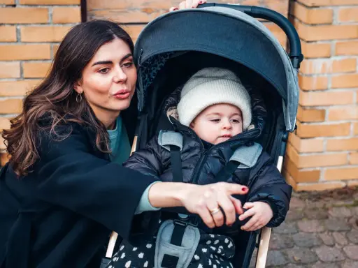 Mama pokazuje coś dziecku w najmniejszej spacerówce. Maluch w czapce i kurtce wygląda ciekawie.