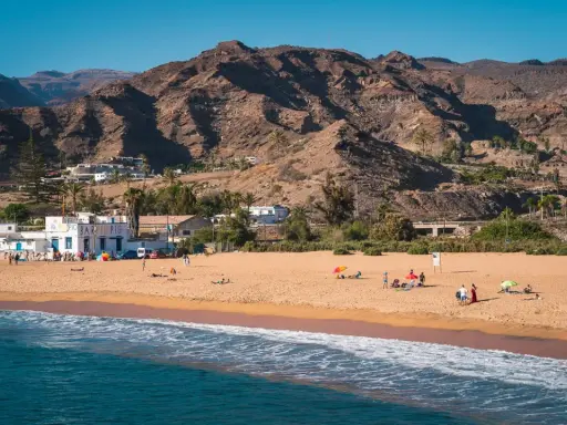 Słoneczne gran canaria plaże z piaszczystym brzegiem, turkusową wodą i górami w tle. Ludzie odpoczywają pod parasolami.