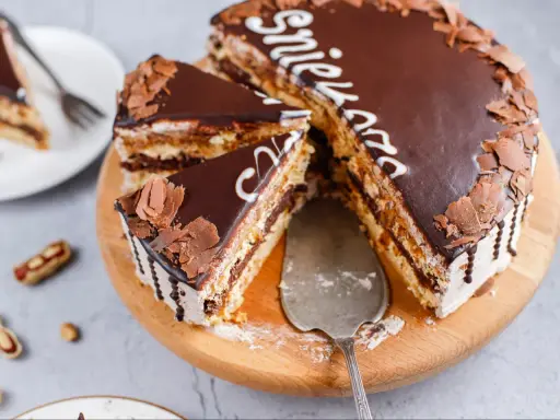 Pyszny tort snickers bez pieczenia, udekorowany czekoladą i wiórkami. Idealny na deser!
