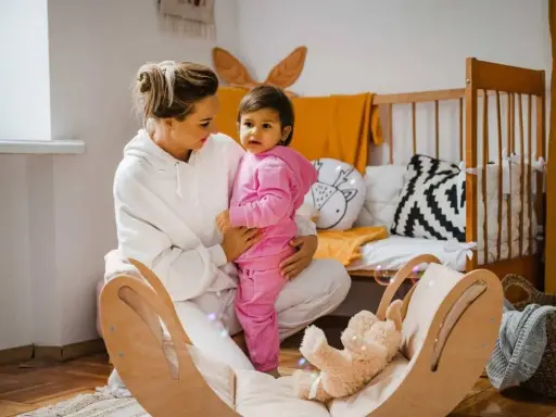 Kobieta w białej bluzie trzyma dziecko w różowym dresie. W tle łóżeczko i drewniana huśtawka z misiem.