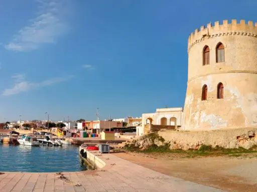Torre Vado, Lecce: un'antica torre di avvistamento domina un porto con barche e case colorate sotto un cielo azzurro.