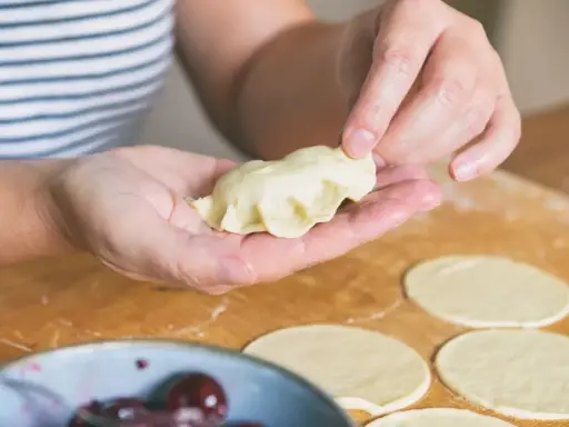 Dłonie formują ciasto na pierogi, tworząc pyszne danie. Na blacie leżą okrągłe placki z ciasta, a obok miseczka z wiśniami.