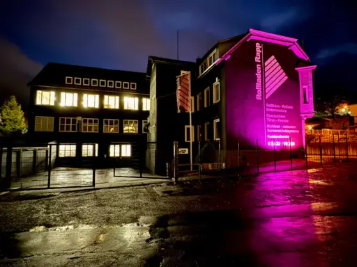 Gebäude mit leuchtenden Fenstern und pinker Beleuchtung. "Rolladen Rapp" wirbt für Nothandkurbel Rolladen, Pflicht für Sicherheit.