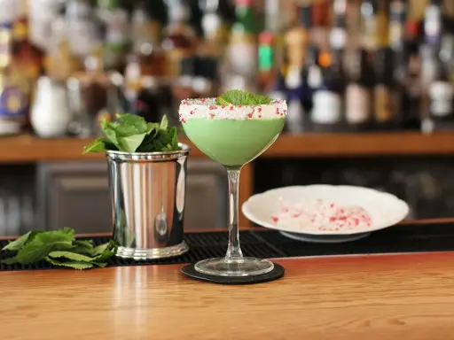 Grön, krämig drink med mynta och krossat strössel på glaskanten. Bredvid står en metallmugg med myntablad och en skål med krossat strössel. Kanske en Amarula drink?