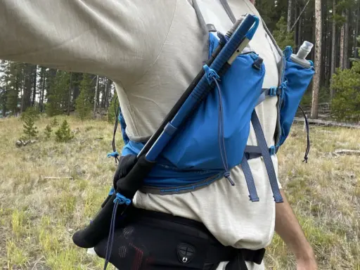 Jak przypiąć kijki do plecaka? Widok z boku na turystę w lesie, z kijami trekkingowymi przymocowanymi do niebieskiego plecaka.