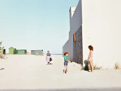 Luigi Ghirri foto: una donna cammina sulla spiaggia, due bambine giocano vicino a un edificio bianco.